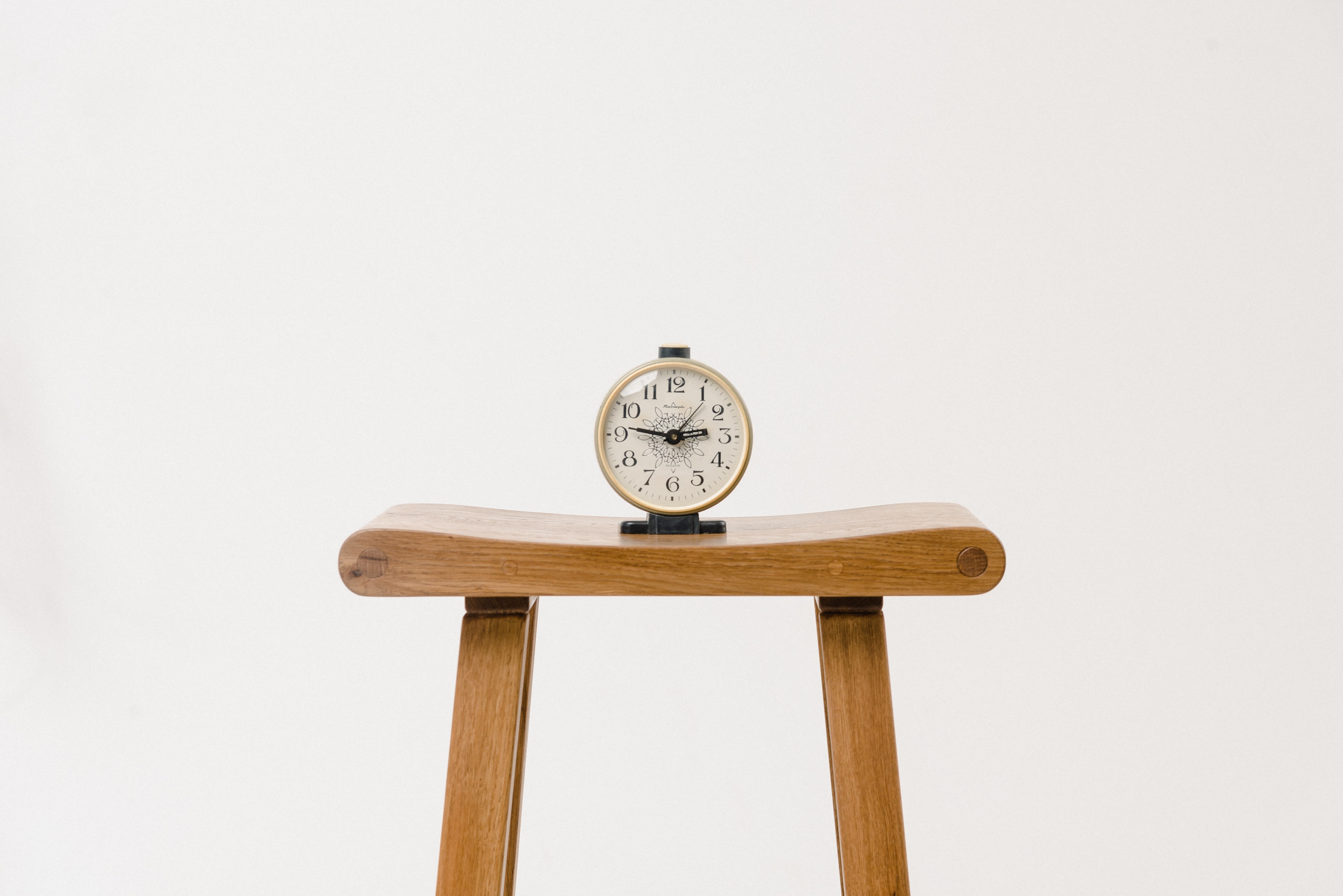 A minimalist photograph featuring a vintage-style alarm clock with a white face and black numerals, placed on a wooden stool against a plain white background. The clock is centered, creating a balanced and clean composition.